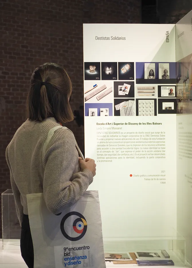 Loida Simarro carrying a "9º Encuentro Bienal Iberoamericana de Diseño - Enseñanza y Diseño" tote bag while observing the "Dentistas Solidarios" project exposition.