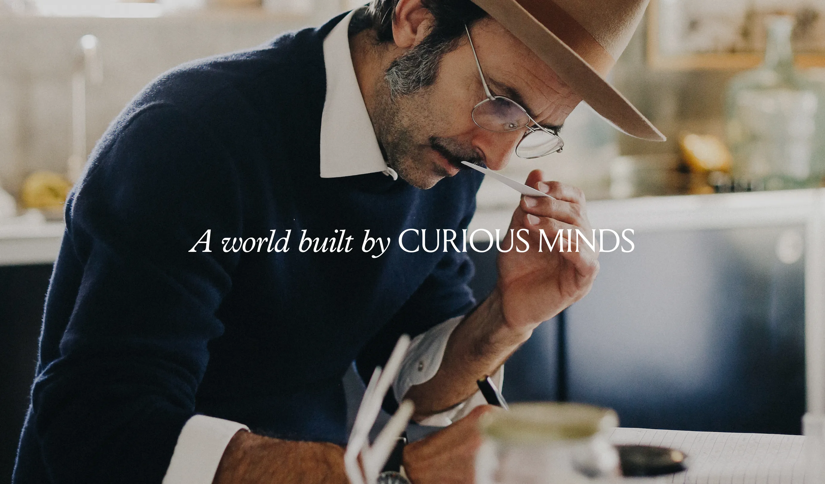 Man with glasses and hat in a blue sweater smelling a fragance while taking notes